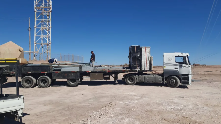 Tractocamión con gabinetes y estructura metálica sobre cama plana junto a una torre de telecomunicaciones